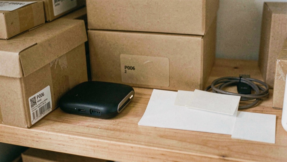 Close documentary photograph of storage boxes, a compact external drive, a cable and blank inventory cards on a wooden shelf, no legible writing, warm daylight. Distinct supporting card P006 2 composition for this editorial entry.