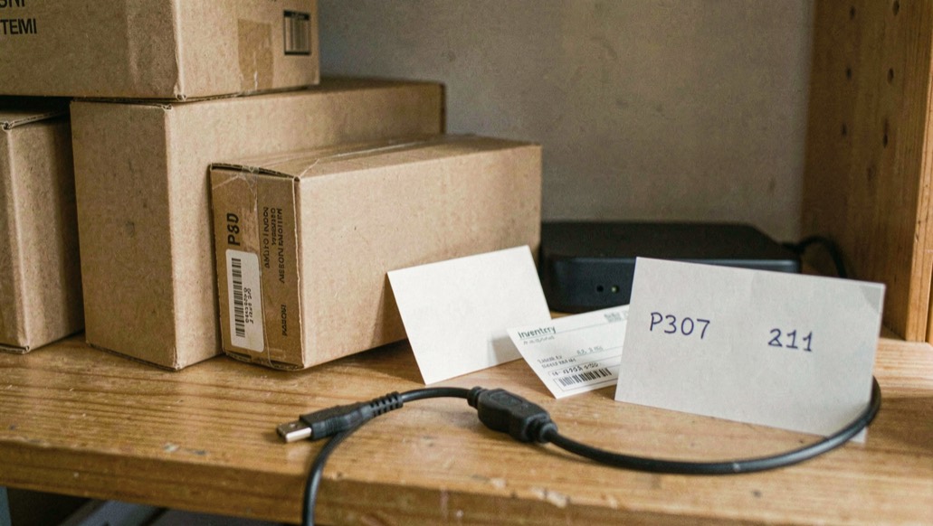 Close documentary photograph of storage boxes, a compact external drive, a cable and blank inventory cards on a wooden shelf, no legible writing, warm daylight. Distinct supporting card P307 2 composition for this editorial entry.