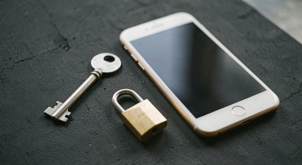 Documentary photograph of a metal key, a phone with a blank dark screen and a small padlock on a charcoal surface, no visible writing, controlled side light. Distinct supporting card P303 3 composition for this editorial entry.