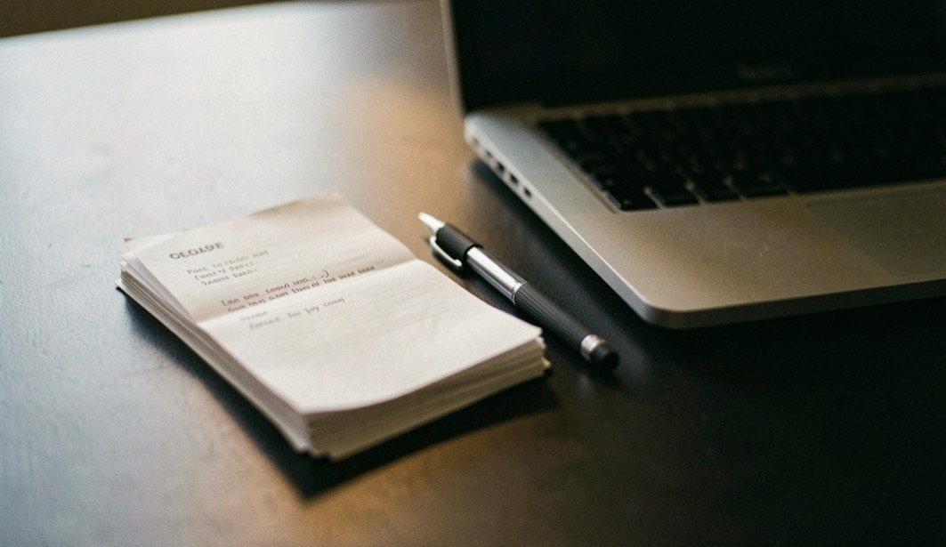 Documentary photograph of a small stack of paper drafts, a mechanical pencil and a compact laptop on a dark desk, no readable words, a single warm pool of light. Distinct supporting card P907 3 composition for this editorial entry.